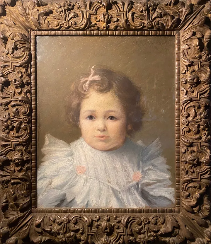 A toddler with curly hair and a pink bow, in an ornate carved frame
