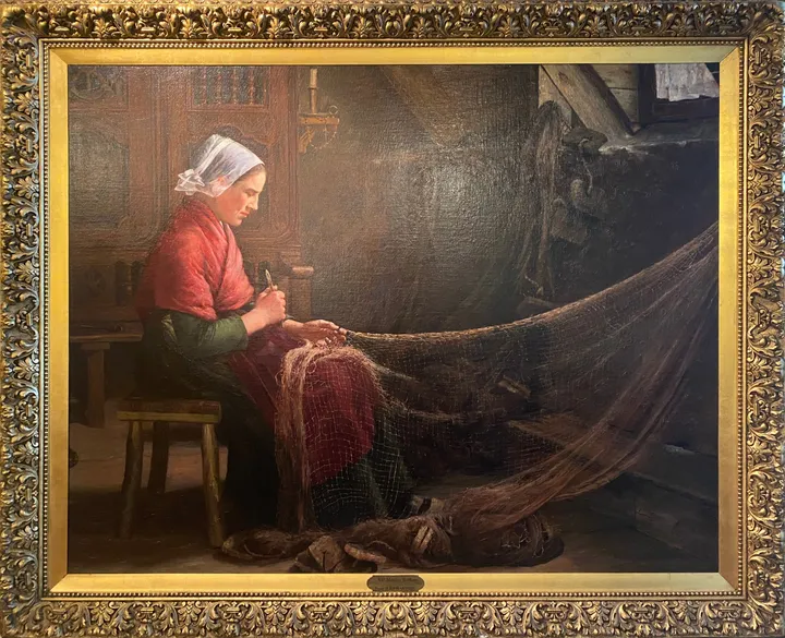 A woman in a white cap and red jacket mends fishing nets in a dim interior