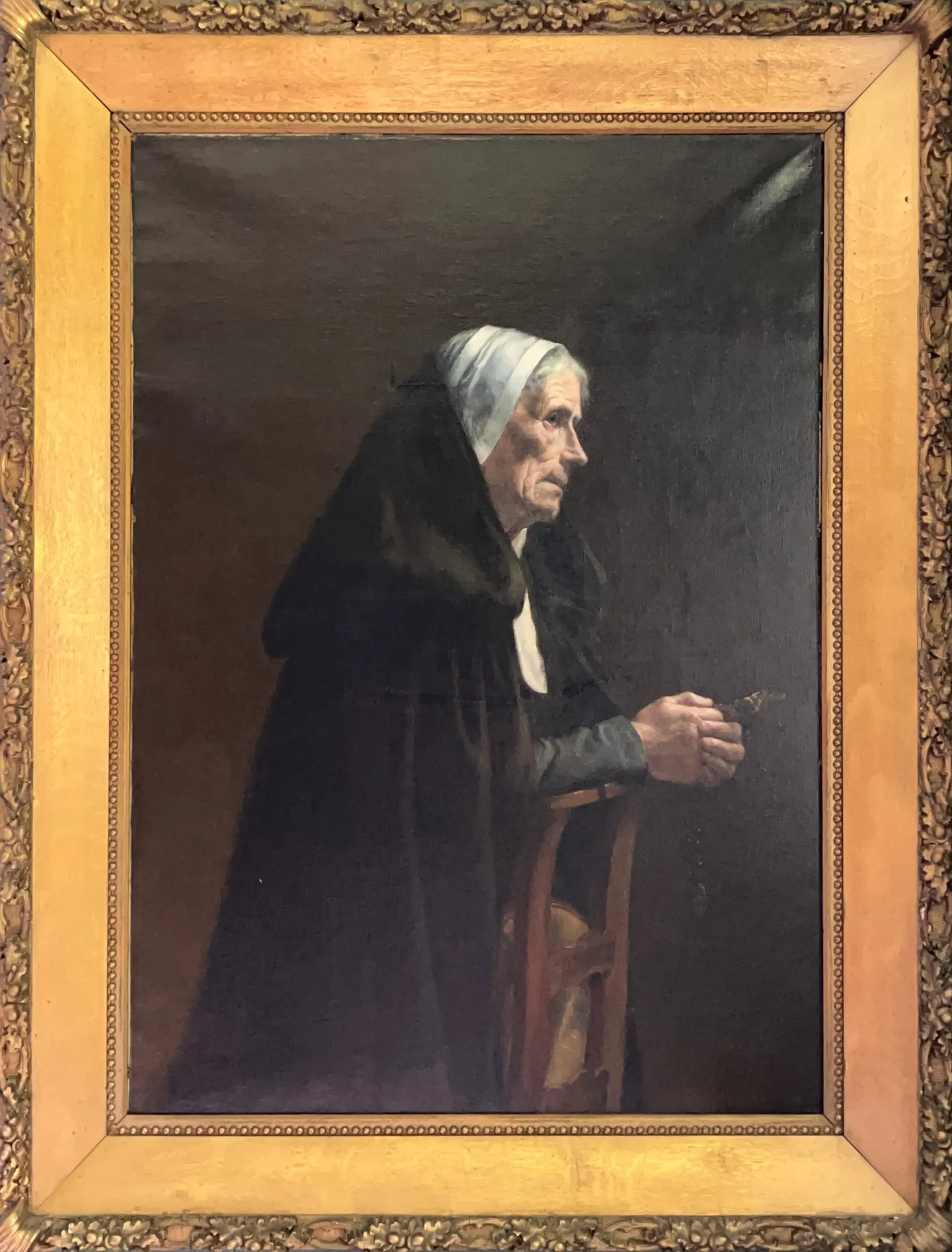 An elderly woman in a black cloak and white cap sits in prayer, holding a rosary