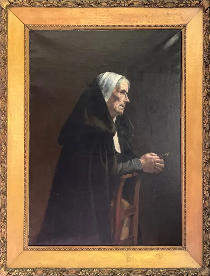 An elderly woman in a black cloak and white cap sits in prayer, holding a rosary